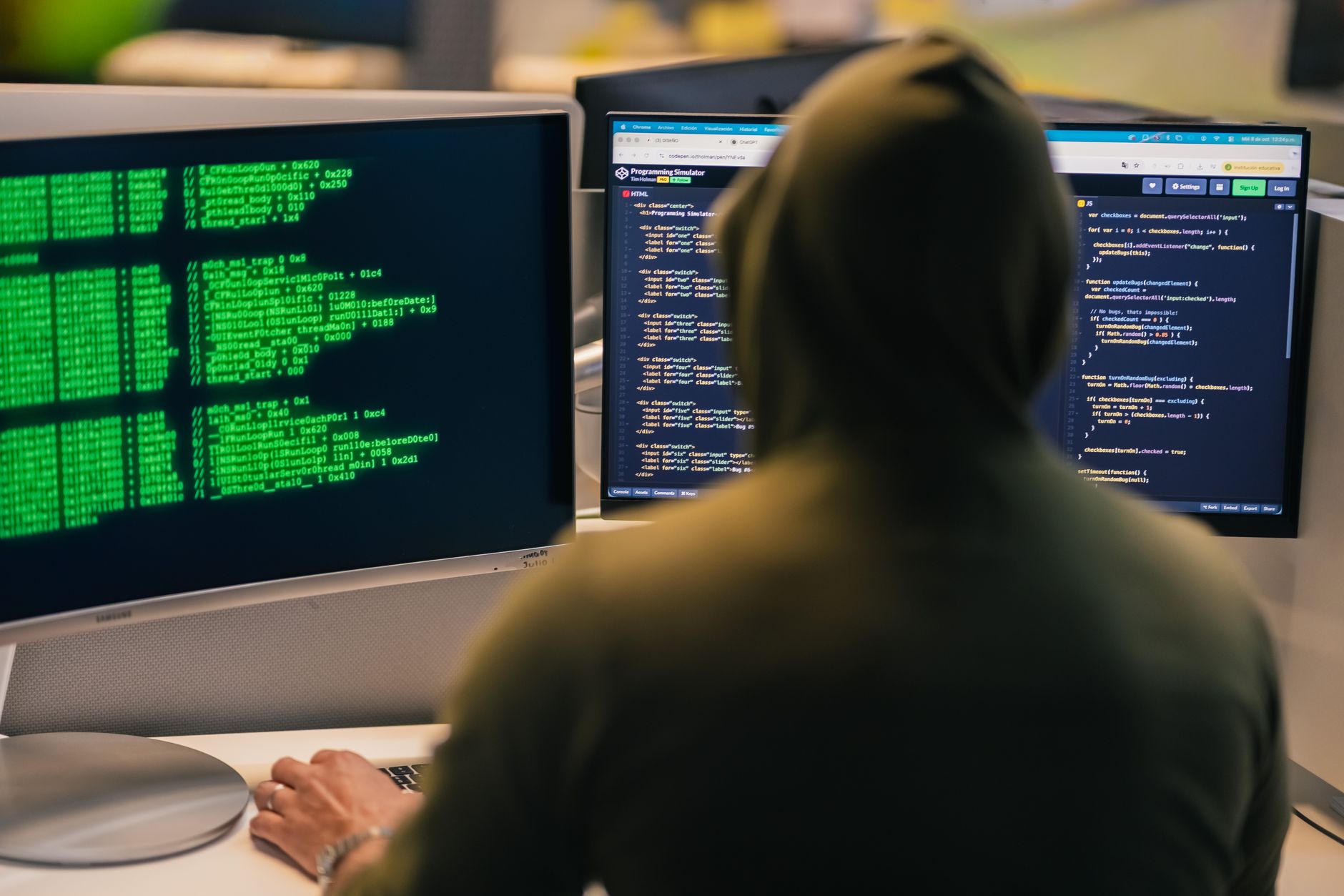 Osoba w kapturze programuje przy dwóch monitorach w ciemnym pomieszczeniu